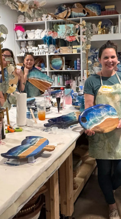 Smiling group of women in an art studio holding blue-green resin “ocean” wooden serving boards at a DIY resin art workshop, paint-covered table with tools, drinks and snacks.