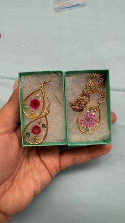 Hand holding small turquoise boxes with gold teardrop handmade resin jewelry — matching earrings and a pendant necklace featuring pink pressed flowers and tiny shell and star charms on a pale blue fabric background.