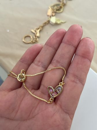 Close-up of a hand holding a delicate gold chain bracelet with a heart-shaped charm filled with pink floral inlay against a soft beige background
