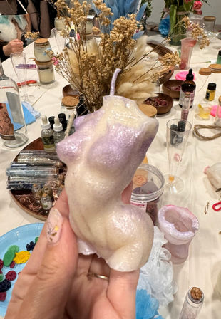 Hand holding a pearlescent pastel lilac female torso candle above a craft table strewn with dried flowers, jars, molds and candle-making supplies — DIY workshop scene.