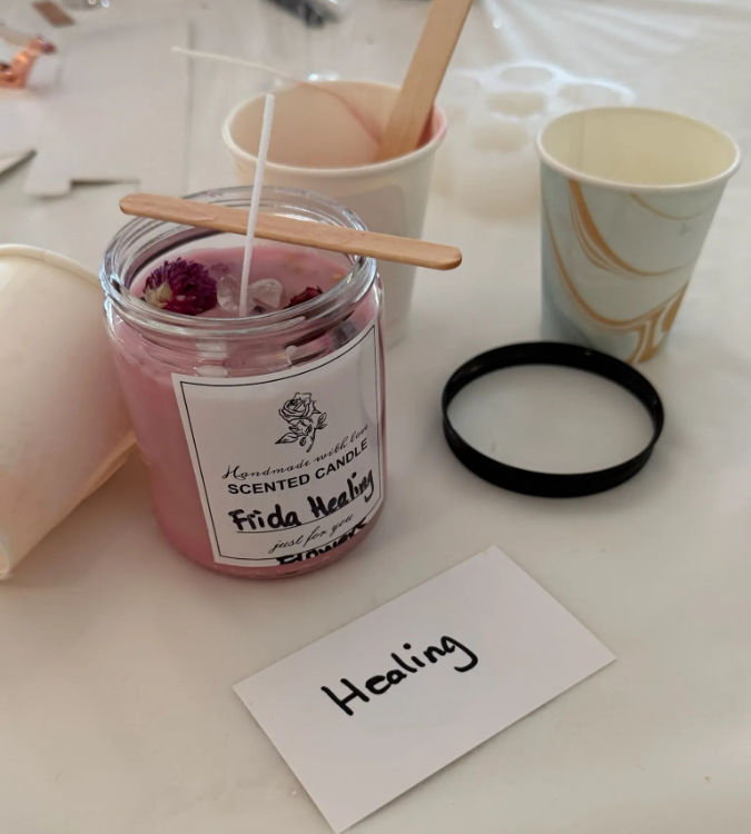 DIY candle-making workshop close-up: handmade pink scented candle in a glass jar topped with dried flowers and crystals, wooden wick holder across the jar, paper cups and a card reading 'Healing' on a craft table.
