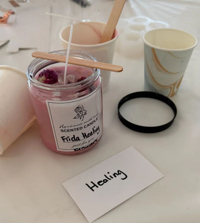 DIY candle-making workshop close-up: handmade pink scented candle in a glass jar topped with dried flowers and crystals, wooden wick holder across the jar, paper cups and a card reading 'Healing' on a craft table.