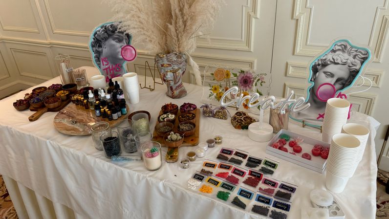 DIY candle and wax-melt workshop table in an elegant white-paneled room, arranged with labeled pigment samples, essential oil bottles, dried botanicals in wooden bowls, silicone molds, paper cups, stirring jars and a playful 'Create' sign.