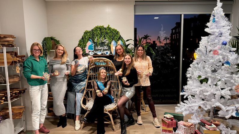 Eight women celebrate indoors at a festive holiday gathering, holding drinks and desserts around a hanging rattan chair, a white decorated Christmas tree and wrapped gifts, with an evening city skyline visible through a window.