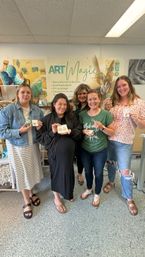 Five women smiling in an indoor art studio holding handmade resin jewelry and craft pieces in front of colorful workshop signage and decor — group photo from a DIY resin art class.