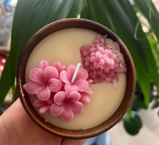 Hand holding a round wooden candle filled with cream wax and pretty pink sculpted wax flowers with crystal accents, decorative floral candle against a blurred indoor plant background