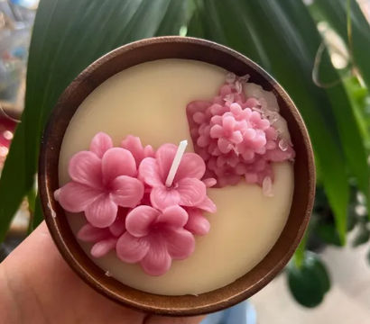 Hand holding a round wooden candle filled with cream wax and pretty pink sculpted wax flowers with crystal accents, decorative floral candle against a blurred indoor plant background