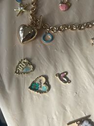 Gold charm bracelet with a shiny heart locket and scattered small heart charms featuring blue enamel, floral accents and a starfish charm on a beige fabric background