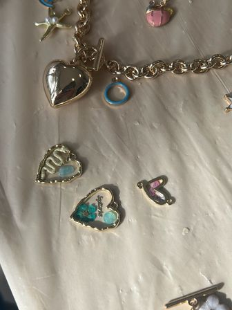 Gold charm bracelet with a shiny heart locket and scattered small heart charms featuring blue enamel, floral accents and a starfish charm on a beige fabric background