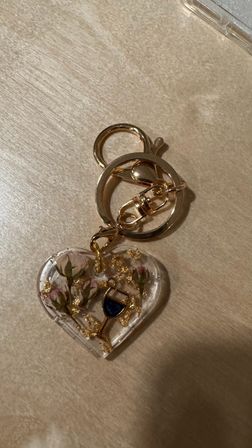 Gold keyring with a clear heart-shaped resin charm holding dried pink rosebuds, gold leaf flakes and a tiny wine-glass charm, resting on a light wood surface.
