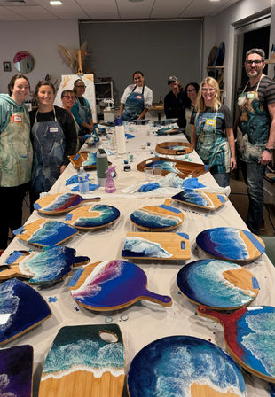 Adults at an indoor resin art workshop posing behind a long table of bamboo boards and paddles decorated with blue ocean wave resin designs, aprons and art supplies visible.