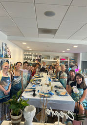 Smiling adults at a paint-night group painting class in a bright indoor art studio, wearing aprons around a long table with canvases, brushes, paint supplies and ceramic shelves in the background.