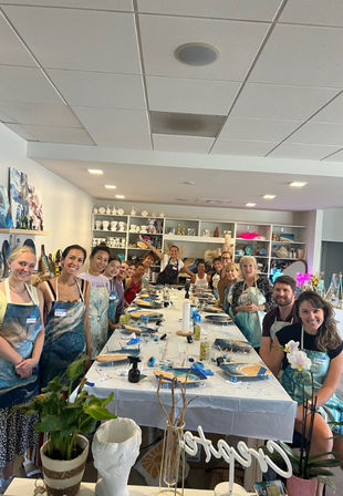 Smiling adults at a paint-night group painting class in a bright indoor art studio, wearing aprons around a long table with canvases, brushes, paint supplies and ceramic shelves in the background.