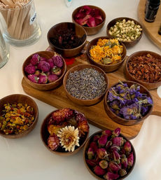 Top-down view of colorful dried botanicals — lavender, rosebuds, calendula, jasmine and purple blooms — arranged in small wooden bowls on a white table.