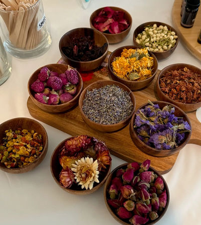 Top-down view of colorful dried botanicals — lavender, rosebuds, calendula, jasmine and purple blooms — arranged in small wooden bowls on a white table.