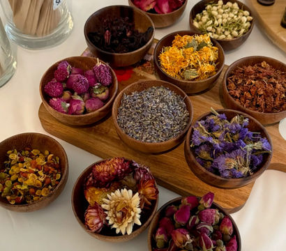 Top-down view of colorful dried botanicals — lavender, rosebuds, calendula, jasmine and purple blooms — arranged in small wooden bowls on a white table.