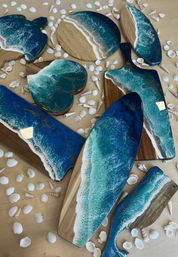Handcrafted wooden serving and charcuterie boards topped with ocean-blue epoxy resin waves and white foam edges, styled on a light surface with scattered seashells for coastal kitchen decor.