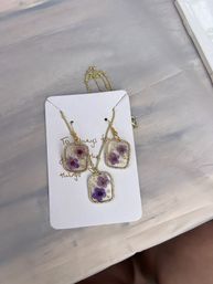 Handmade resin floral jewelry set with pressed purple flowers and gold flakes — pendant necklace and matching dangle earrings in gold-tone frames on a white display card, flatlay on a pale marble surface.