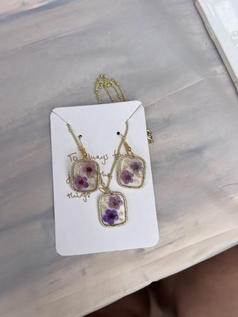 Handmade resin floral jewelry set with pressed purple flowers and gold flakes — pendant necklace and matching dangle earrings in gold-tone frames on a white display card, flatlay on a pale marble surface.