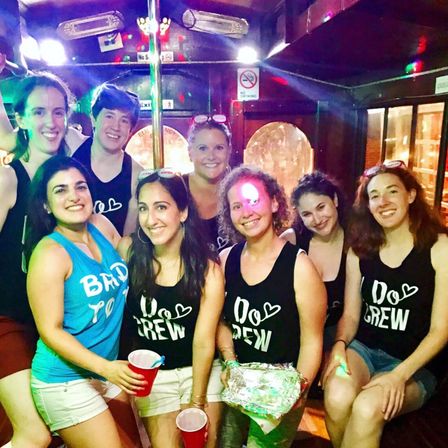 Bachelorette party group of eight women wearing matching Do Crew black tanks and one in a blue Bride-to-be top, holding red cups and smiling inside a party bus/trolley with a center pole, wood interior and colorful disco lights.