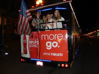 Nighttime downtown trolley back deck with a group of smiling women holding drinks, an American flag, and a large red energy-drink advertisement on the rear, lit by streetlights and passing traffic.