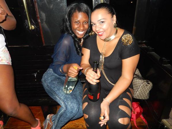 Two women smiling in a nightclub booth celebrating with champagne bottles, wearing party outfits and accessories with a handbag nearby.