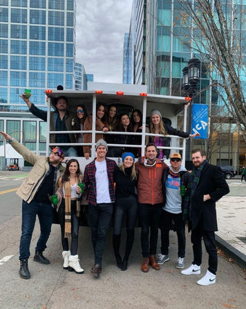 Group of friends smiling and holding drinks while posing on and around a white open-sided party truck on a downtown street with modern glass office towers