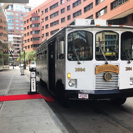 White vintage party trolley parked on a downtown city curb next to a red carpet and valet signs, with brick office buildings and glass storefronts in the background.