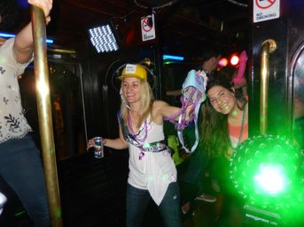 Bachelorette party on a neon-lit party bus: two women smiling and dancing, one wearing a "bride-to-be" hard hat and beads while holding a can and a playful bra, colorful LED and green disco lights in the background.