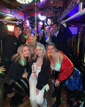 Group of smiling young adults posing inside a crowded party bus with a central pole, colorful disco lights and a birthday sash, night-out celebration vibe.