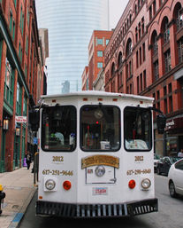White tour trolley parked on a narrow Boston street flanked by red brick historic buildings and a glass skyscraper in the background.