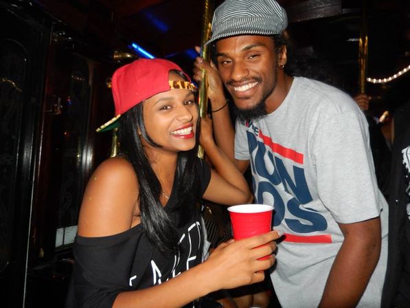 Cheerful young adults at an indoor party or nightclub — woman in a backwards red cap holding a red solo cup and smiling, man in a striped cap and graphic tee leaning by a brass pole.