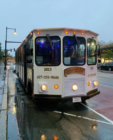 White vintage-style party trolley parked on a rainy downtown street at dusk, amber headlights and blue interior lights glowing, reflections in puddles with a streetlamp and storefronts in the background.