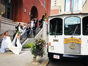 Joyful wedding party in tuxes and light-blue bridesmaids descending red-brick building steps to board a white party trolley on a downtown city sidewalk — bride with flowing veil and bouquet.