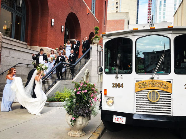 Joyful wedding party in tuxes and light-blue bridesmaids descending red-brick building steps to board a white party trolley on a downtown city sidewalk — bride with flowing veil and bouquet.