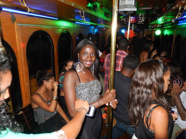 Smiling woman holding a brass pole on a crowded neon-lit party trolley, young adults dancing and chatting in a lively nightlife scene.