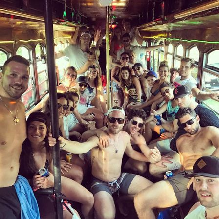 Large group of young adults partying inside a crowded vintage trolley during daytime, many in swimwear and sunglasses holding drinks and smiling