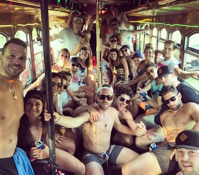 Large group of young adults partying inside a crowded vintage trolley during daytime, many in swimwear and sunglasses holding drinks and smiling