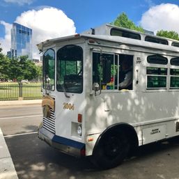 White vintage trolley bus numbered 2004 parked on a city street beside a leafy urban park, glass skyscraper and blue sky in the downtown skyline