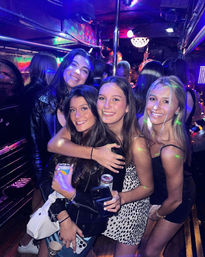Four smiling friends posing together inside a crowded party bus/nightclub with colorful disco lights, holding drinks and phones, enjoying a night out.