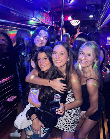 Four smiling friends posing together inside a crowded party bus/nightclub with colorful disco lights, holding drinks and phones, enjoying a night out.
