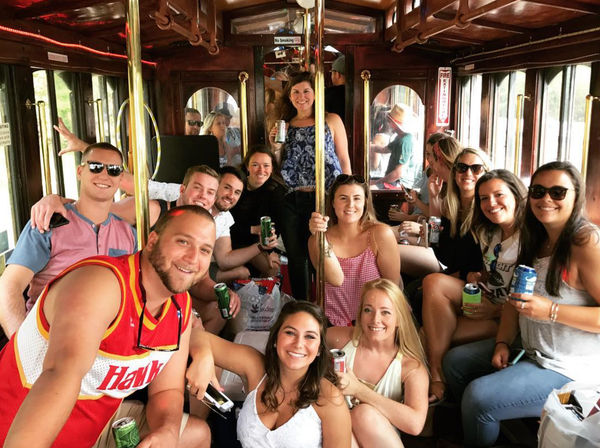 Smiling group of friends holding canned drinks aboard a vintage wooden trolley during a lively daytime city tour