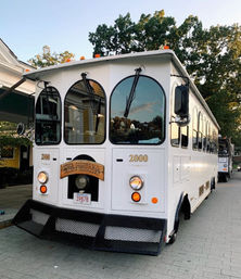 White vintage-style party trolley with arched windows and front sign parked on a tree-lined paved street near storefronts at dusk.