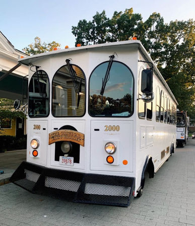 White vintage-style party trolley with arched windows and front sign parked on a tree-lined paved street near storefronts at dusk.