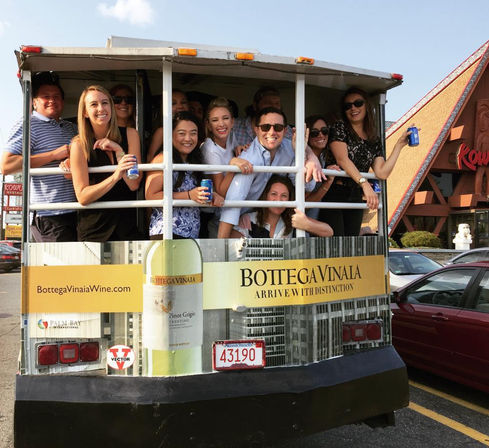Smiling group of friends crowded on an open-air party trolley holding canned drinks, parked in a sunny lot outside a tiki-style restaurant.
