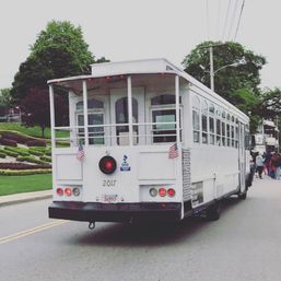 White vintage-style trolley with small American flags rolling down a tree-lined suburban street past landscaped gardens and pedestrians