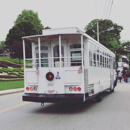 White vintage-style trolley with small American flags rolling down a tree-lined suburban street past landscaped gardens and pedestrians