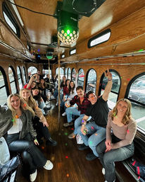 Cheerful group of friends partying on a wood-paneled city trolley, disco lights and brass poles overhead, holding drinks and smiling beside arched windows showing parked cars on a downtown street.