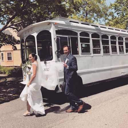 Wedding couple — bride in a white gown holding a bouquet and groom in a navy suit walking past a vintage white trolley on a sunny tree-lined neighborhood street.