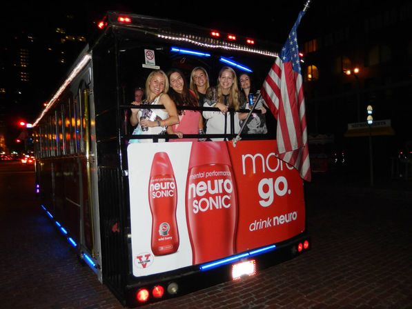 Smiling group of women on the back of an illuminated party trolley at night on a downtown brick street, American flag and bold red beverage ad on the rear panel